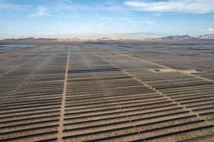 An aerial view of a vast expanse of rows of solar panels extending nearly as far as the eye can see through the desert toward ridges of arid mountains in the distance.