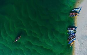 Aerial view looking directly down at a collection of multi-colored canoes sitting in the water near a beach. The water is a green color with patterns of waves clearly visible.