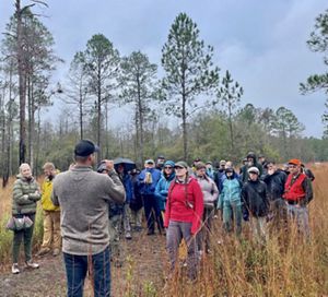 A group of people wearing rain gear stand outdoors and listen to a speaker.