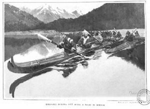 An old, black-and-white photo features several men rowing a long boat in the water, with mountains behind them. 