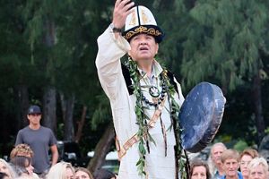 A man wearing white regalia and holding a drum participates in a ceremony.