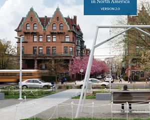 Several people enjoy the swings in a city park. Traffic drives by and rowhouses face the park across the street.