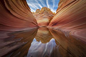 Vista a nivel del suelo de paredes rocosas anaranjadas en el desierto con una poza de agua en el centro que refleja la formación rocosa anaranjada situada a distancia.