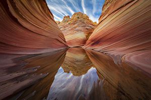  Ground-level view of orange 'striped' desert rock walls with a pool of water in the center that reflects the standing orange rock formation in the distance.