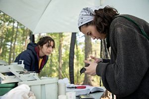 A woman holds a small bird while standing at a table under an open sided tent. Another women standing across from her records notes.