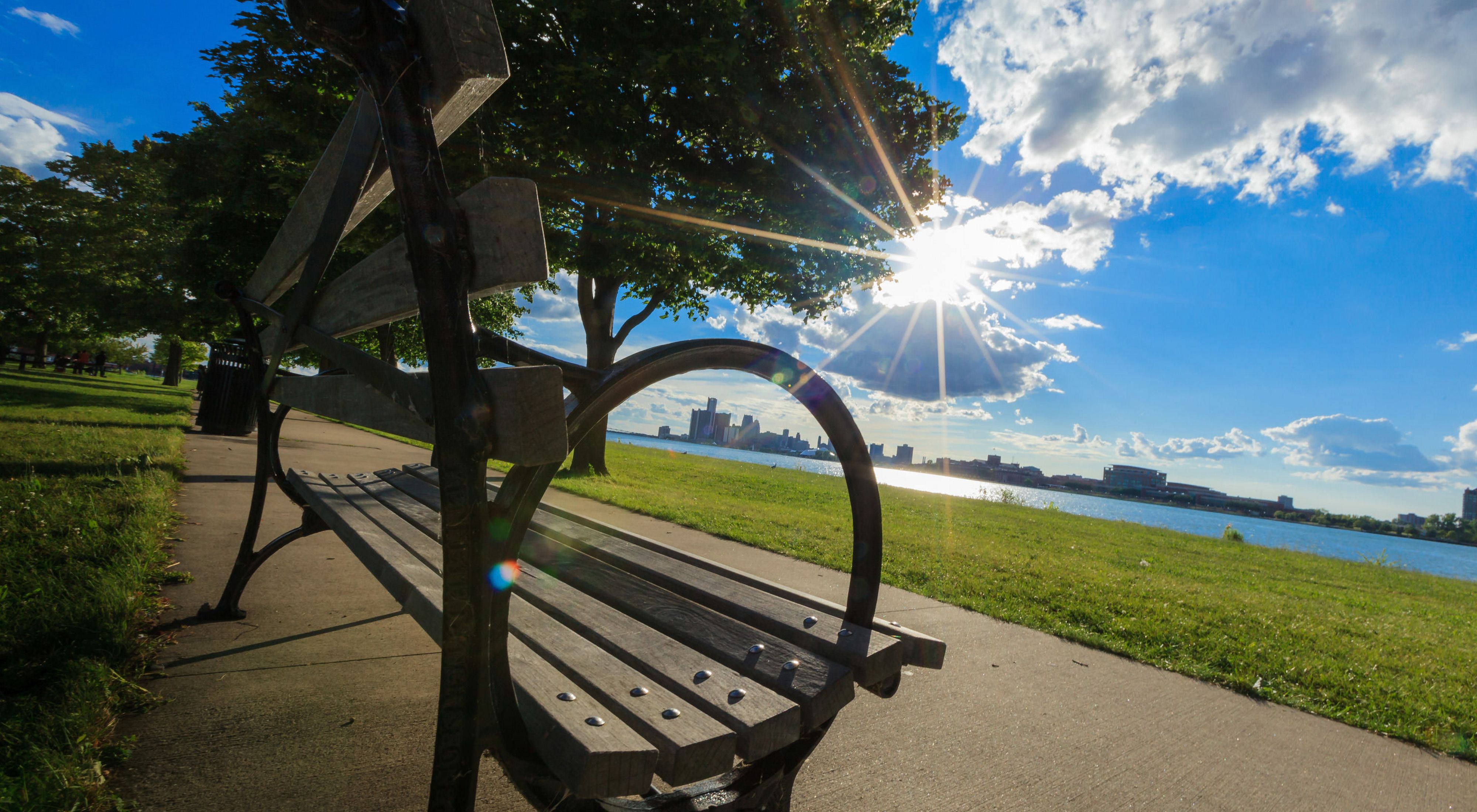 A cast iron and wood park bench along a sidewalk, facing the Detroit River with sun shining through clouds and the city skyline on the opposite shore.