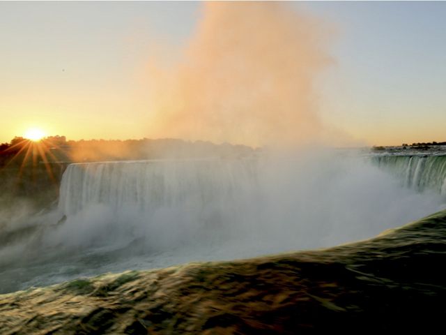 A cloud of mist rises above Niagara Falls as the sun sets over trees in the distance.