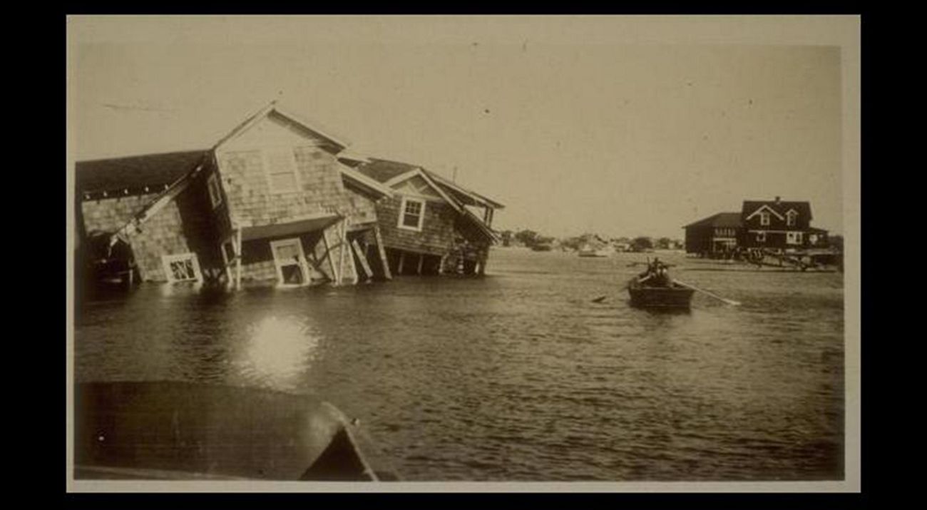 Vintage sepia toned photo showing severe flooding after a storm. A person rows a boat between houses that have been knocked off their foundations and are laying tipped into the deep water.