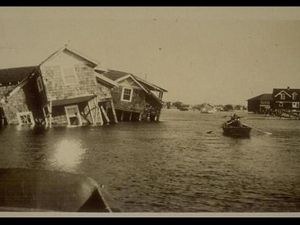 Vintage sepia toned photo showing severe flooding after a storm. A person rows a boat between houses that have been knocked off their foundations and are laying tipped into the deep water.