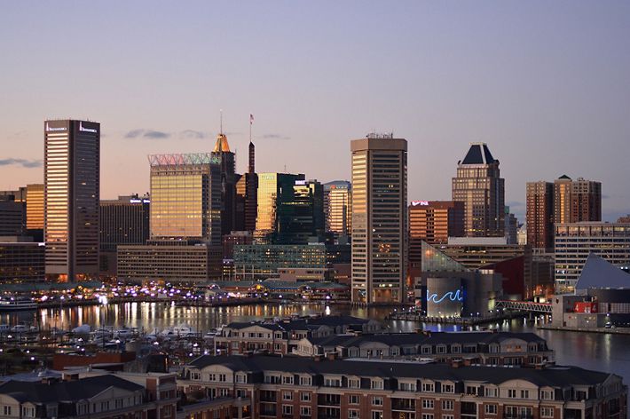 The sun sets over the Baltimore skyline. Tall building dominate the background with low rowhouses lining the foreground.