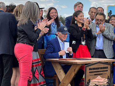 President Biden sits at a wooden desk with individuals standing around him, clapping.