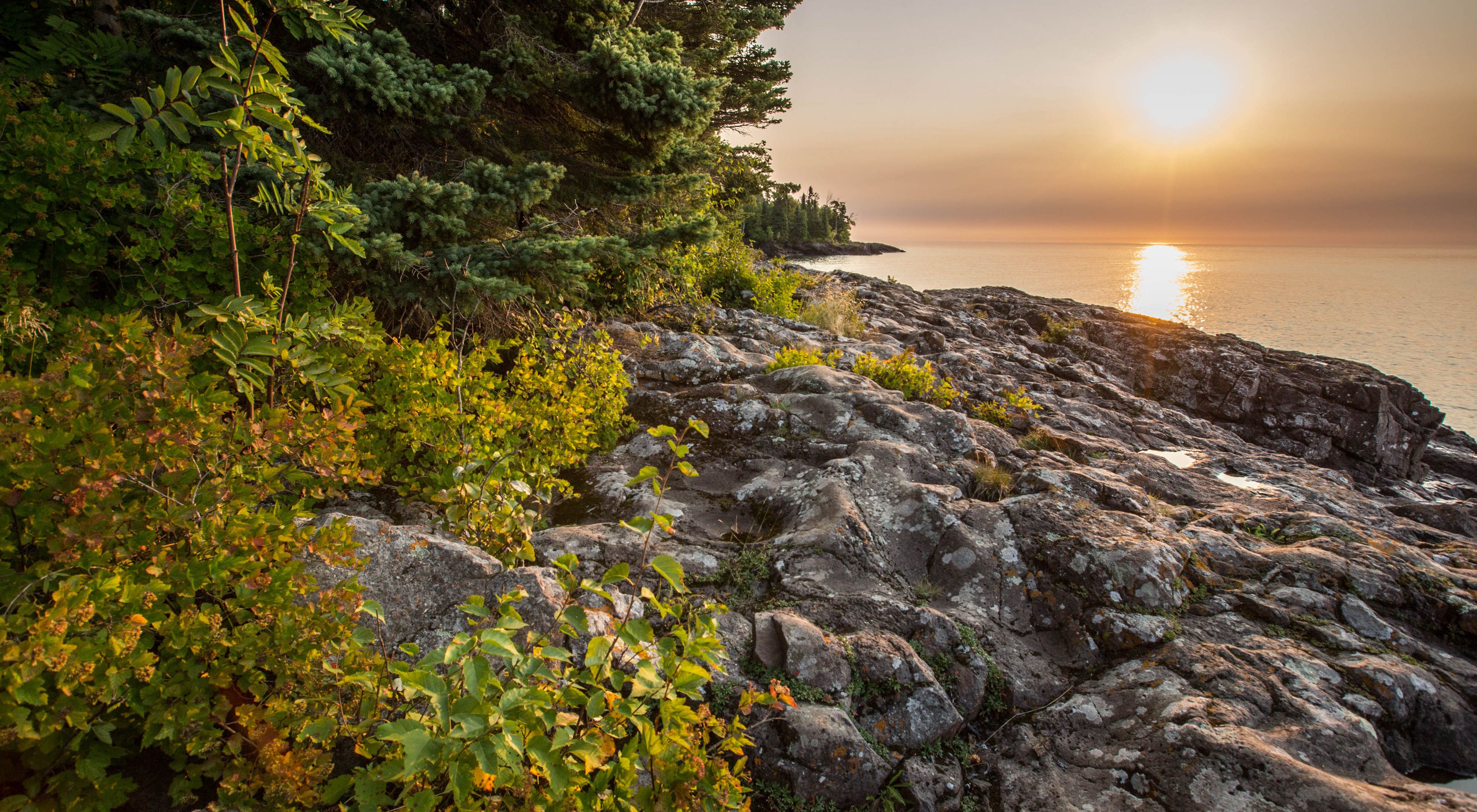 The glow of sunset over a tranquil lake with rocky, rugged, forested shoreline in the foreground.