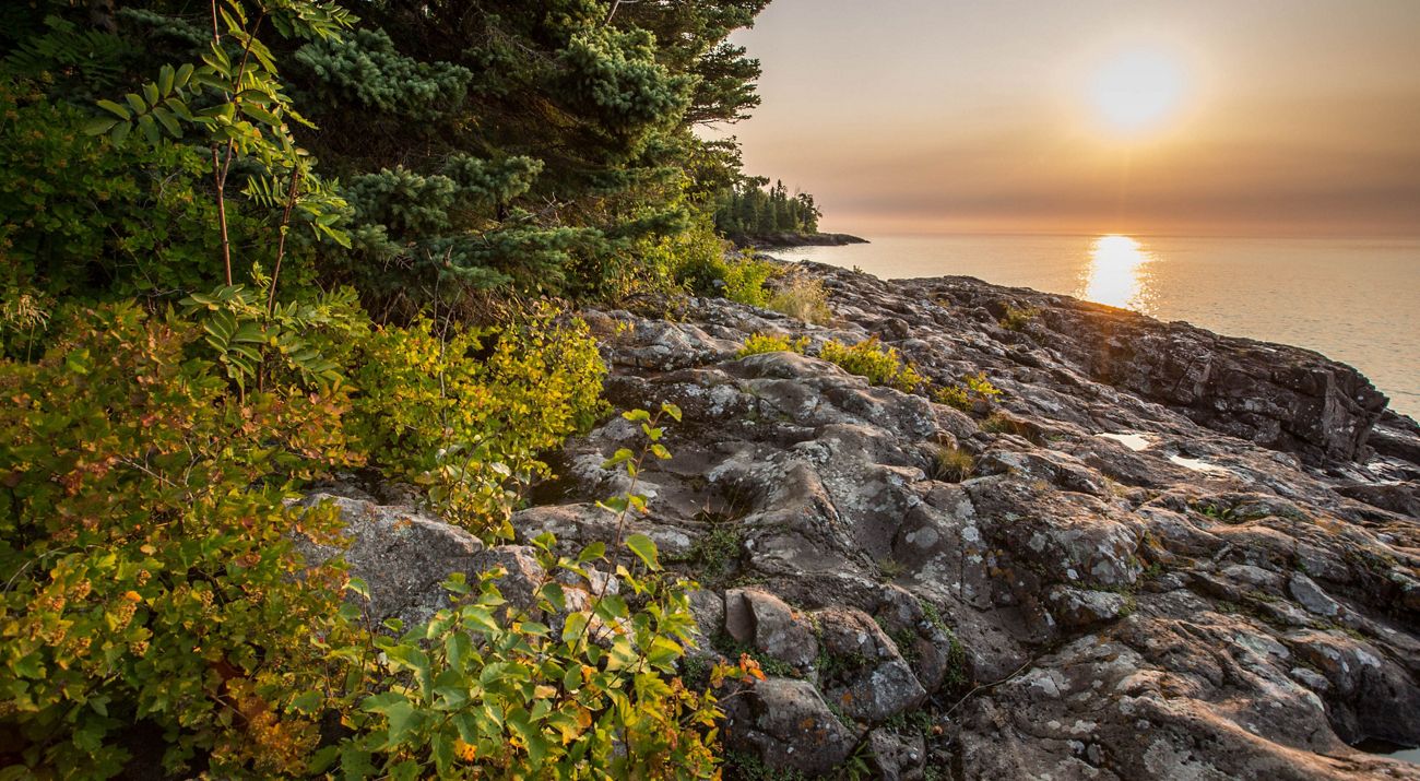 Dense forest along rocky shoreline with sun setting over a calm lake.