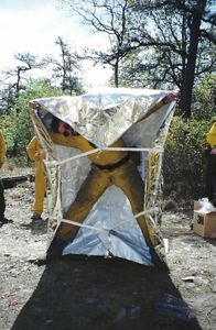 An old photograph of a person in yellow fire gear holding a silver fire blanket over their head. 
