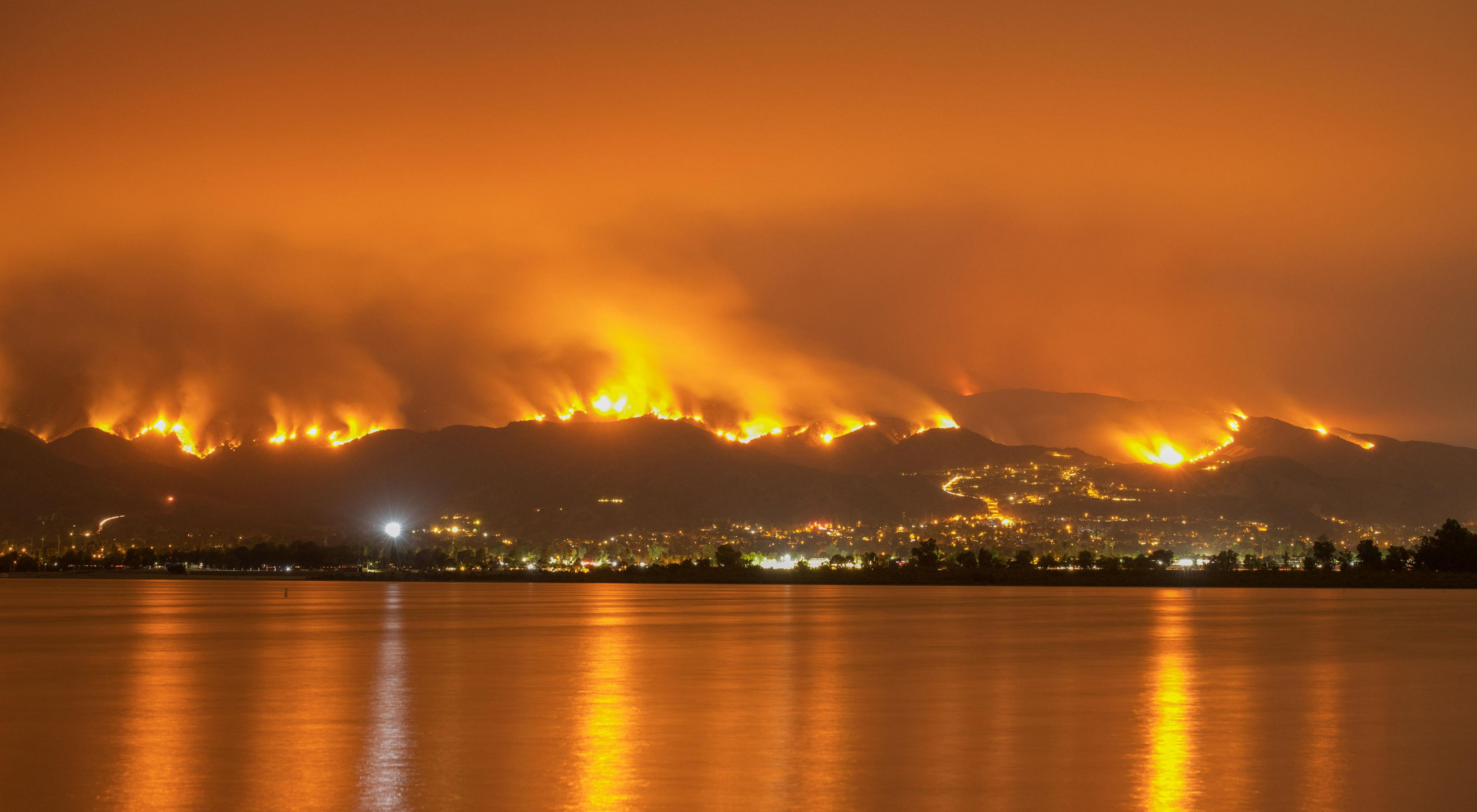 One of the 2018 California wildfires. A glowing orange sky with water in the foreground and flames rising over hills in the background.