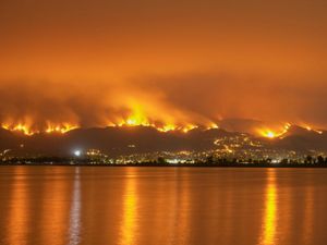 A view across water at hillsides burning with orange flames at night.