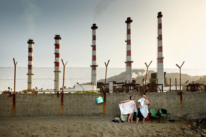 Two people in shorts and t-shirts, carrying towels, stand on brown sand against a concrete wall as the red-and-white smokestacks of a factory are seen in the distance.
