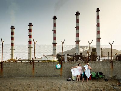 Two people stand in the sand, holding towels in front of their bodies. Behind them is a cement and chainlink wall separating the beach from a large factory.
