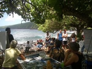A group of people on a shady beach gather around a map and look towards a speaker. 