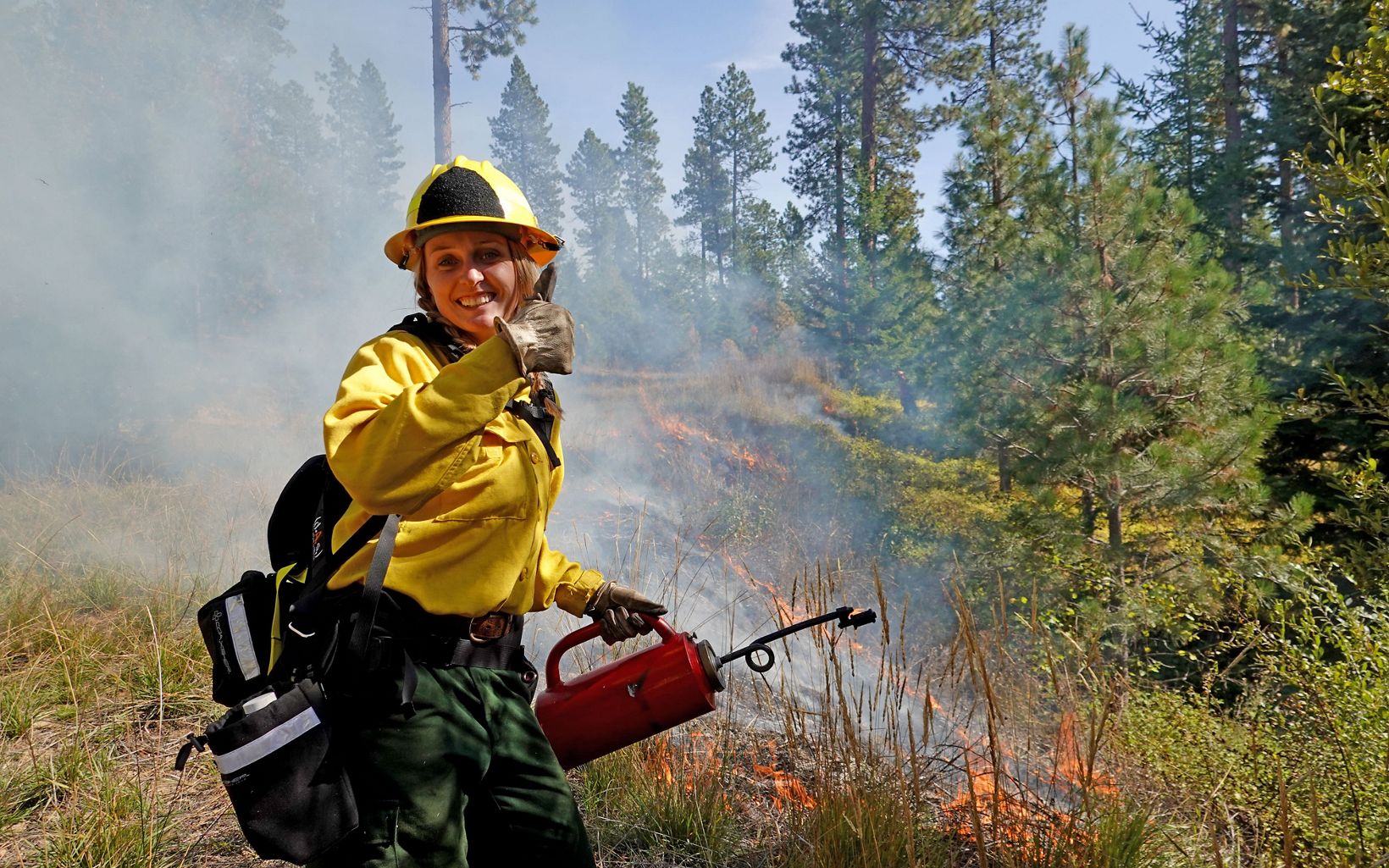 A fire fighter grins and gives a thumbs up while holding a drip torch. Fire burns the grasses.