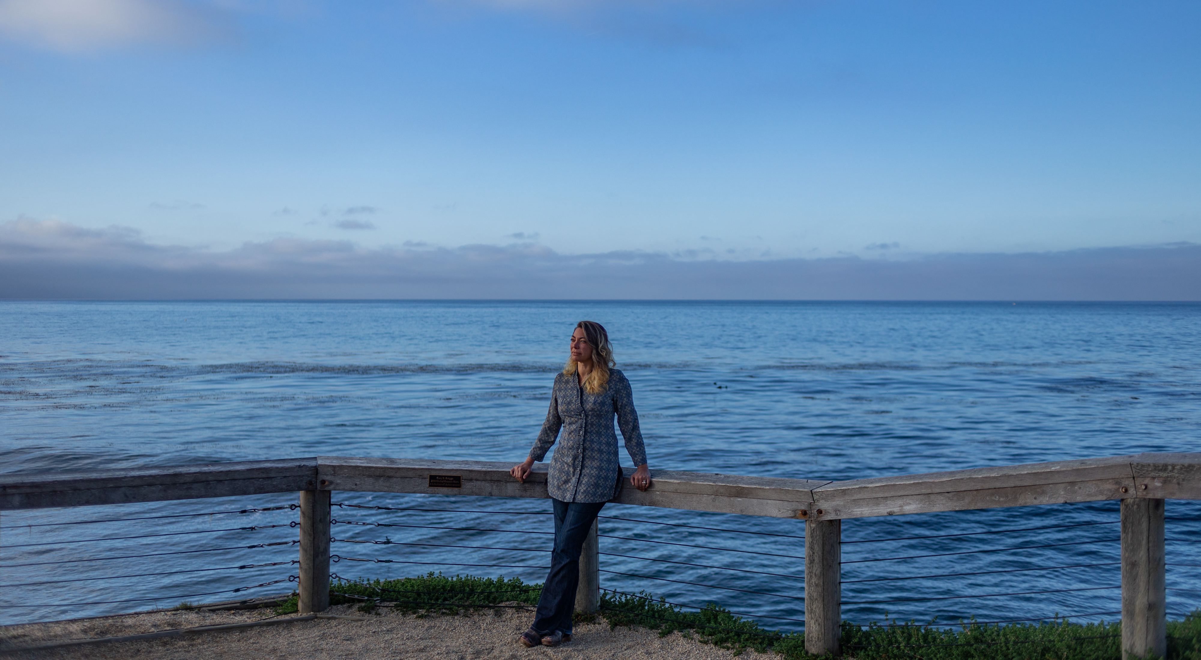 TNC scientist Heather Tallis leans against a railing facing the camera, with a vast blue Pacific Ocean horizon behind her.