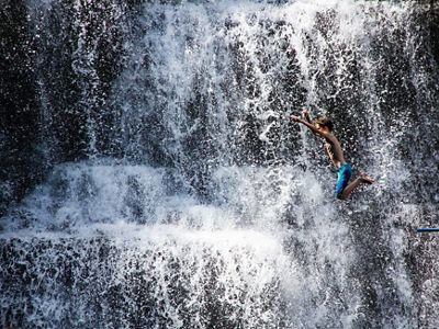 A boy jumps into the water with a large waterfall in the background.