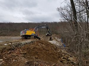 A large yellow backhoe sits on top of an earthen dam. A large breach has been opened in the area in front of the machine. The bucket is full of dirt and rocks.