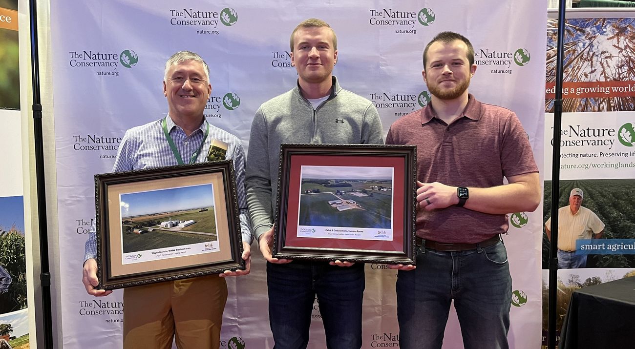 Three smiling people holding framed photos in front of a backdrop.
