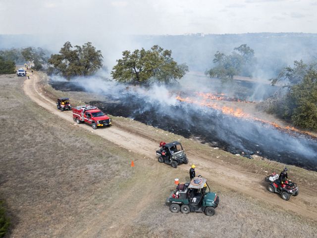 A group of fire practitioners stand by four ATVs and two fire trucks on a road serving as a fire break, as orange flames spread across shrubland habitat.