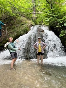Two individuals stand in shallow water near a waterfall.