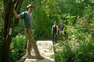Three people gather around a boardwalk trail in a forest.