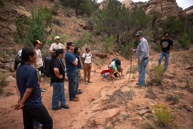 Several college students stand outside in a red-rock landscape and listen to an adult speaking to them.