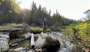 A man stands on a large rock in the middle of a flowing creek.
