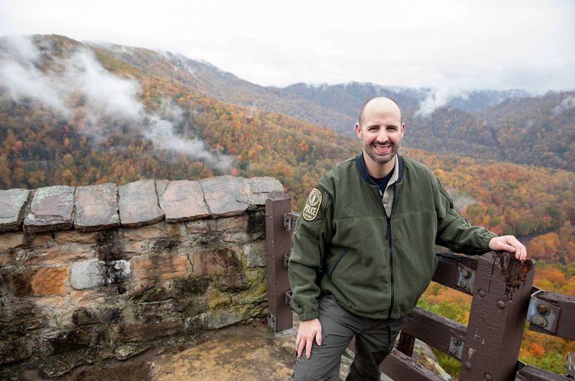 A smiling man stands at a mountain overlook. Mountain ridges and valleys roll away to the horizon behind him.