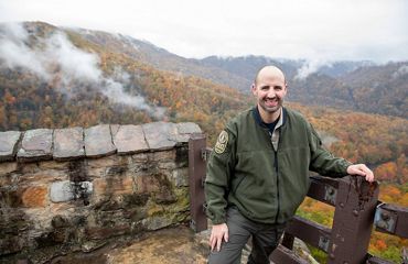 A smiling man poses next to a fence surrounded by a colorful mountain valley.