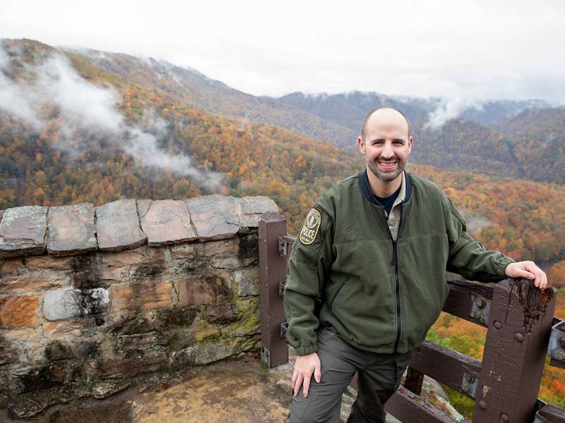 A smiling man stands at a mountain overlook. Mountain ridges and valleys roll away to the horizon behind him.