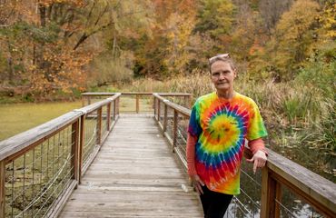 A woman wearing a colorful tie die poses on a boardwalk surrounded by forest.