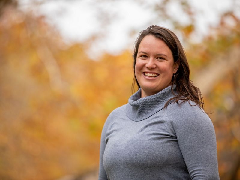Candid portrait of Idalina Walker standing in front of orange and yellow, out of focus leaves.