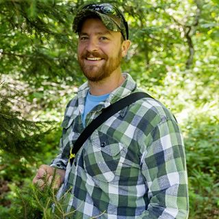 Headshot of Nick Proctor smiling and standing among bright green trees.