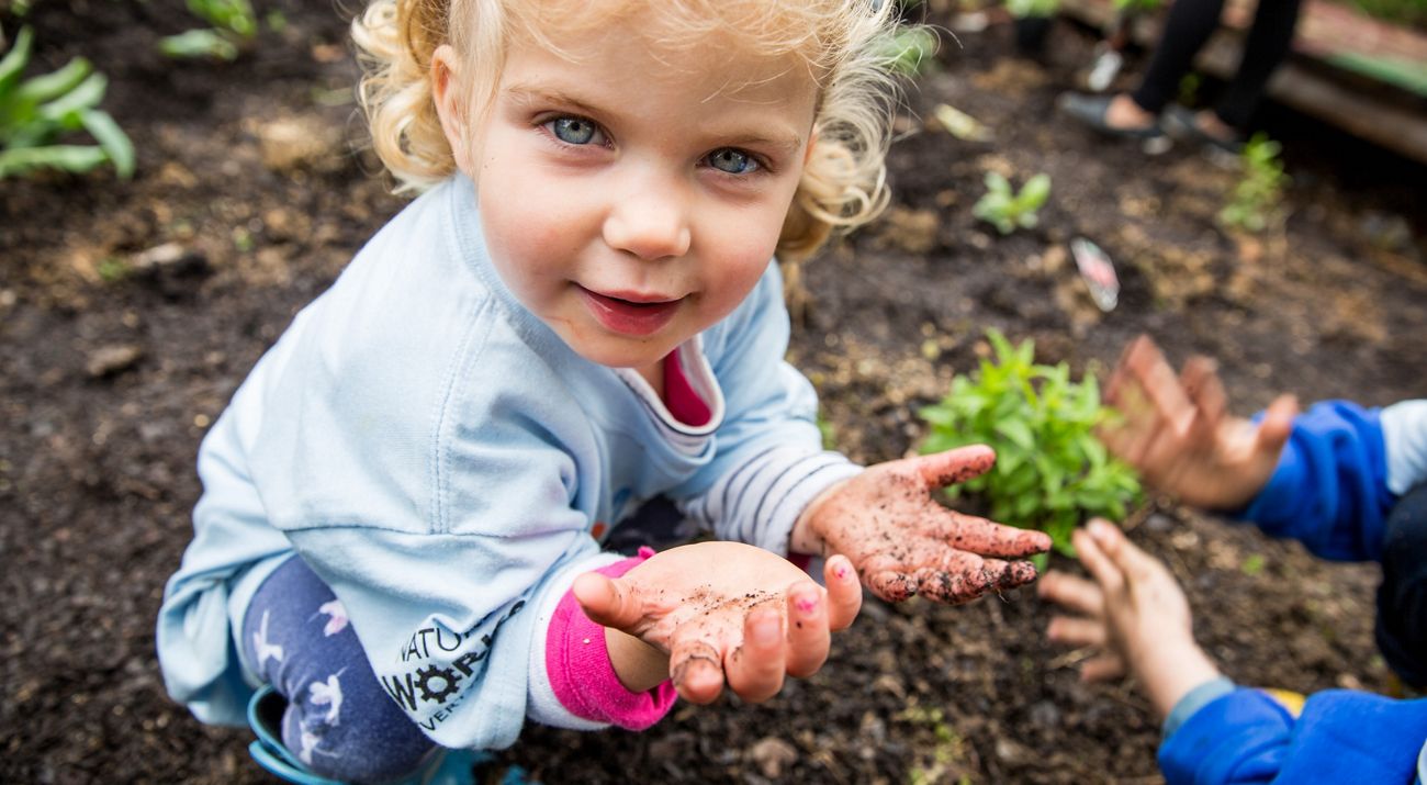 A little girl crouches in a garden, looking at the camera. Her open hands are covered in fine soil.
