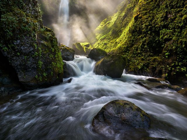 Sunlight illuminates a waterfall flowing through a moss-covered gorge