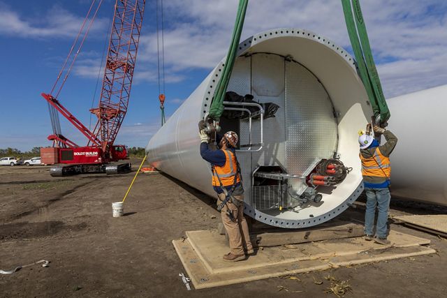 Photo of two construction workers setting straps on the base of an industrial wind turbine in preparations for installation. 