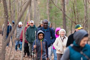 A large group of people walk along a path through a forest. The diverse group is bundled up against the early spring chill.