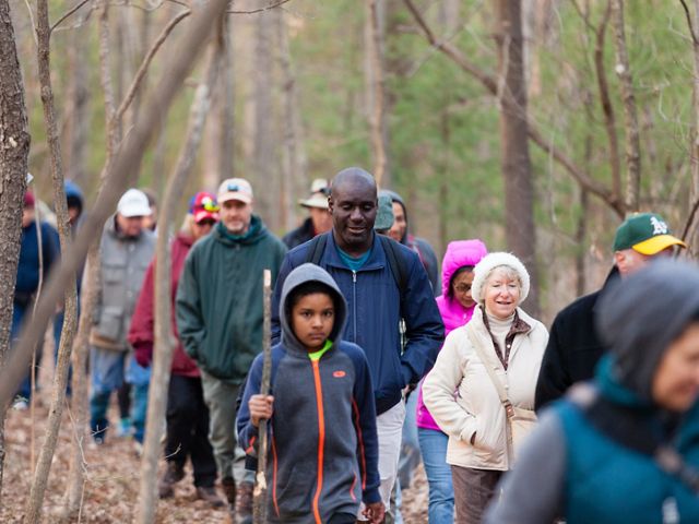 A large group of people walk along a path through a forest. The diverse group is bundled up against the early spring chill.