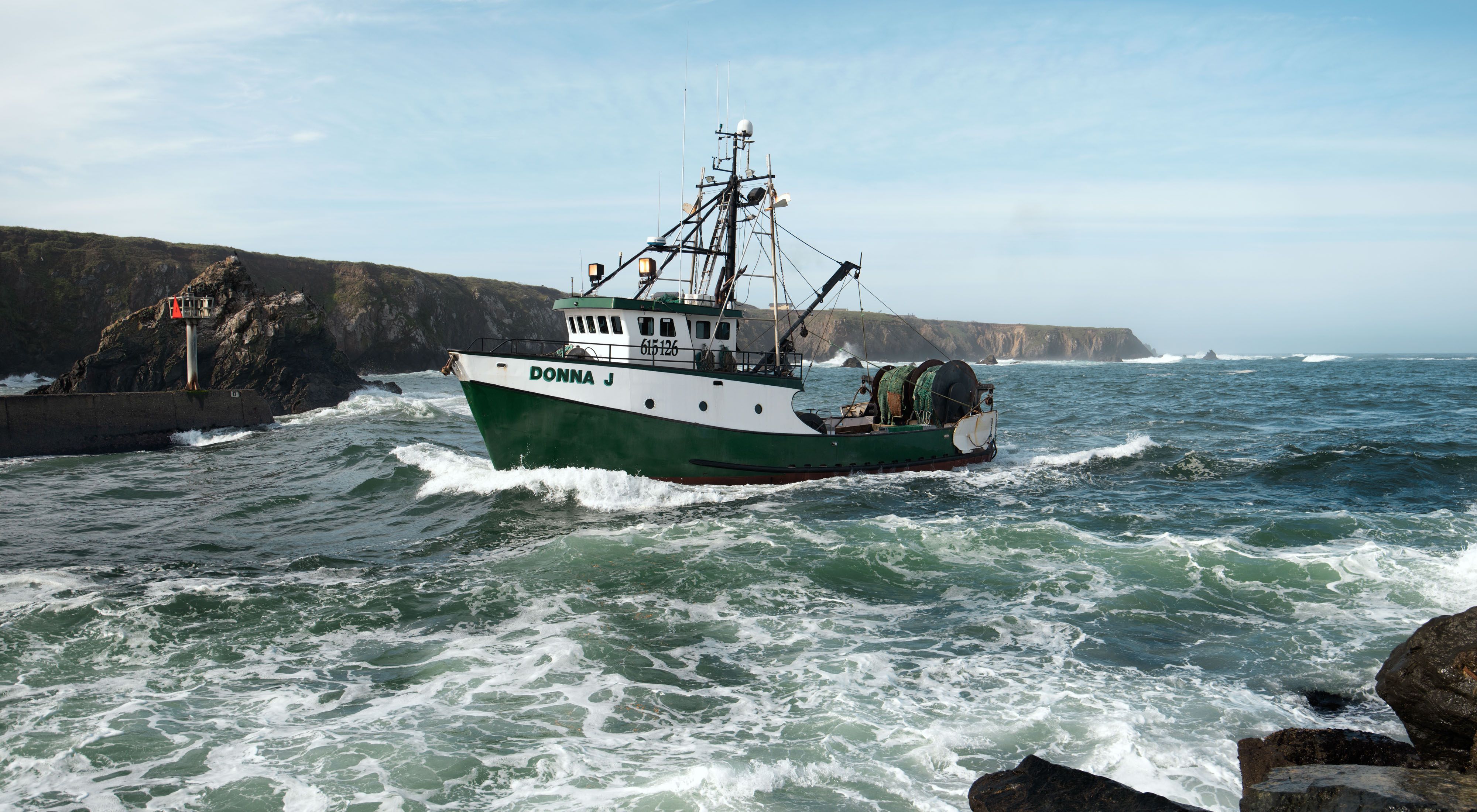 A green-and-white fishing boat named the Donna J motors into a rocky harbor on choppy seas.