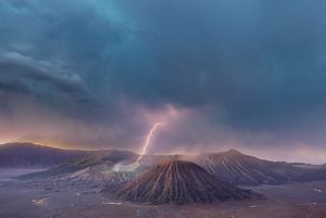 A lightning bolt flashes above a volcanic landscape under thick, heavy clouds.