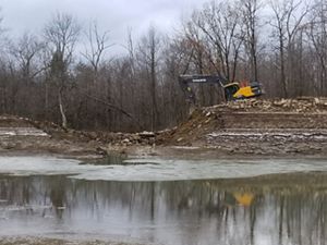 A large yellow backhoe sits on top of an earthen dam. The large breach in the dam reveals the stone that faces the back side of the dam. A few inches of water remain in the reservoir.