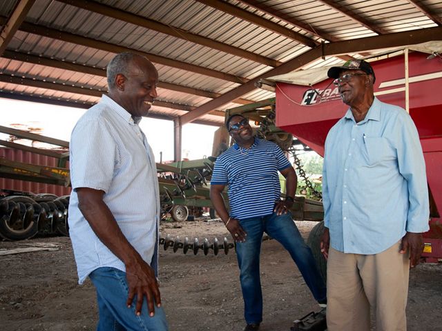 Three men stand near farm equipment in a a large barn.