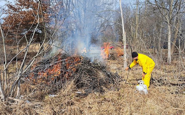 A land manager in protective gear lights a pile burn in the foreground. In the background, other piles of debris are lit by additional managers. 