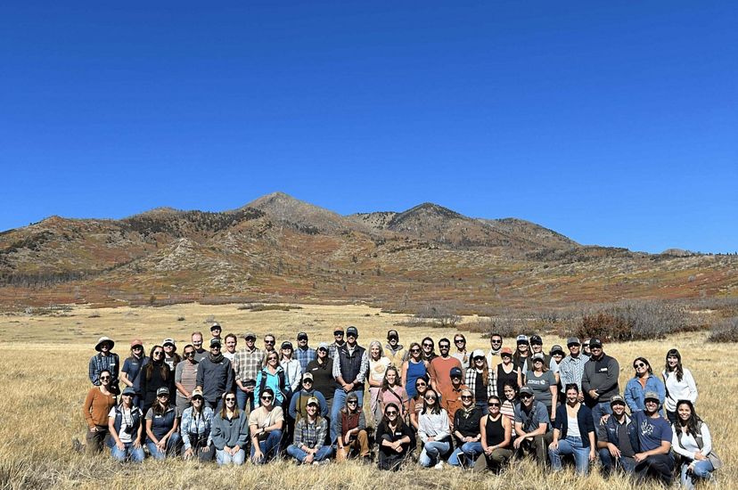 A group of 50 people smiling in a big grass field with mountains in the background.